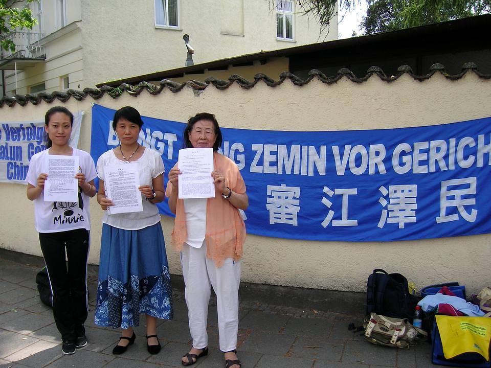 Tri praktikantice Falun Dafa iz Minhena prikazuju svoje krivične prijave protiv Jiang Zemina zbog progona Falun Dafa. Vidjevši transparent: "Izvedite Jiang Zemina pred lice pravde" na kineskom i njemačkom, <a href="http://en.minghui.org/html/articles/2015/7/26/151739.html">jedna žena se zaustavila i pitala šta se dešava</a>. Nakon što je čula da su praktikantice bile zlostavljane zbog njihovog vjerovanja u Istinitost-Dobrodušnost-Toleranciju, rekla je: "Našem društvu trebaju ljudi kao što ste vi [praktikanti]. Molim vas, nastavite s vašim akcijama!"